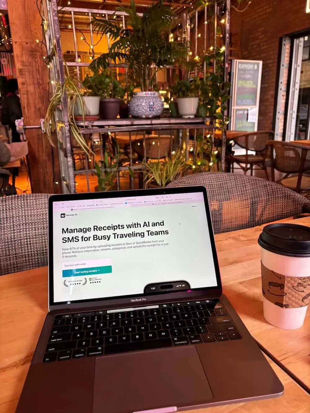 A warm café interior filled with hanging plants, string lights, and wooden furniture. In the foreground, a MacBook Pro rests on a wooden table, open to the Receipt AI website displaying the headline “Manage Receipts with AI and SMS for Busy Traveling Teams.” Next to the laptop sits a takeaway coffee cup with a cardboard sleeve. The soft lighting, greenery, and quiet atmosphere reflect a calm moment of focus — a builder working quietly, bringing their vision to life when no one’s watching.
