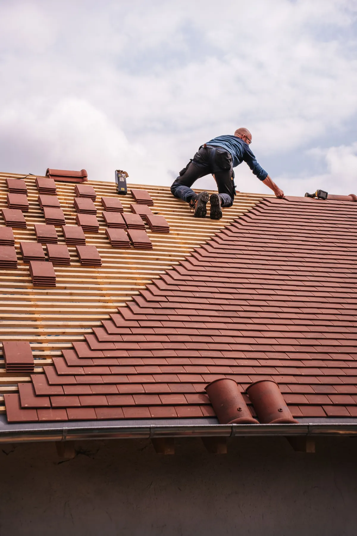 A roofer carefully installs shingles on a new residential roof under a cloudy sky