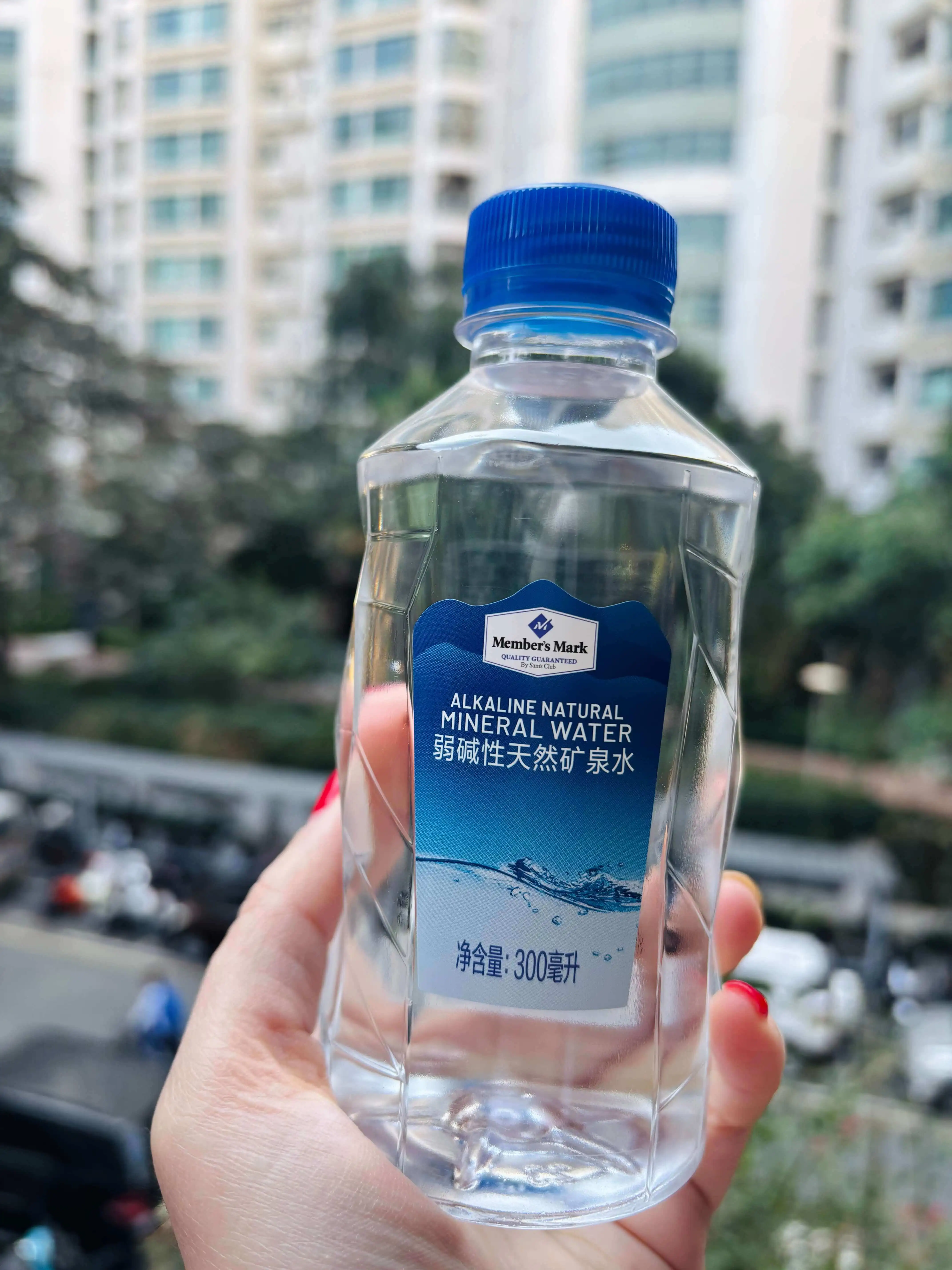 Close-up of a small clear plastic bottle of Members Mark alkaline natural mineral water being held outdoors. The bottle has a blue cap and a blue label with English and Chinese text, and contains 300 mL. The background is blurred high-rise apartment buildings, trees, and parked cars.
